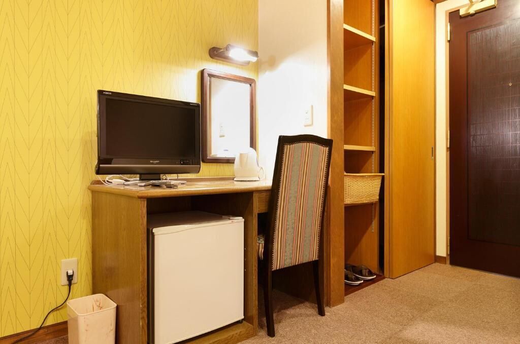 A compact hotel room with a desk, a television, a small refrigerator, a chair, a mirror, and a wardrobe.