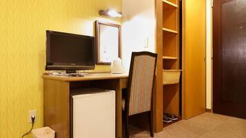 A compact hotel room with a desk, a television, a small refrigerator, a chair, a mirror, and a wardrobe.