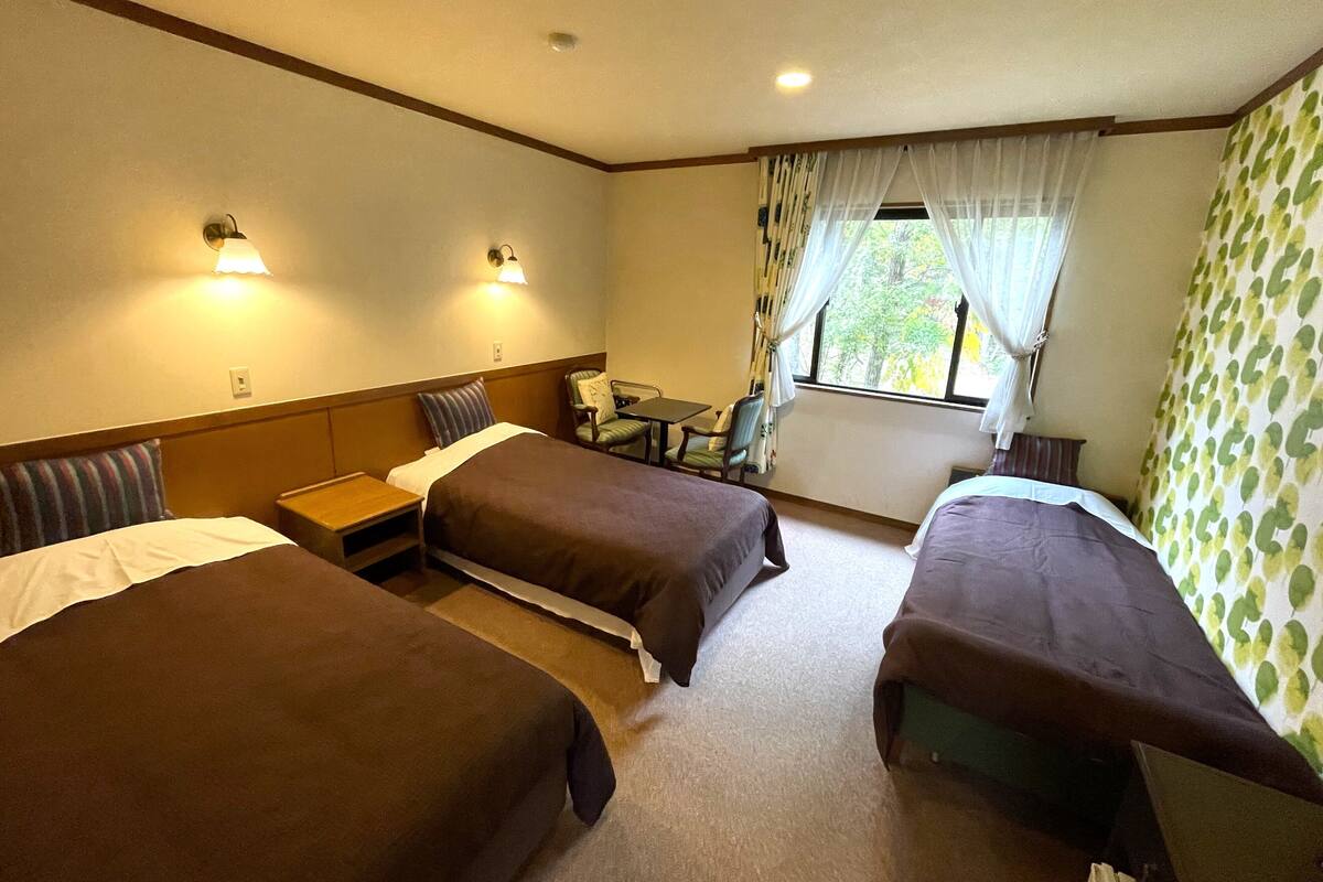 A hotel room with two single beds, a small table, a chair, and a window with curtains.