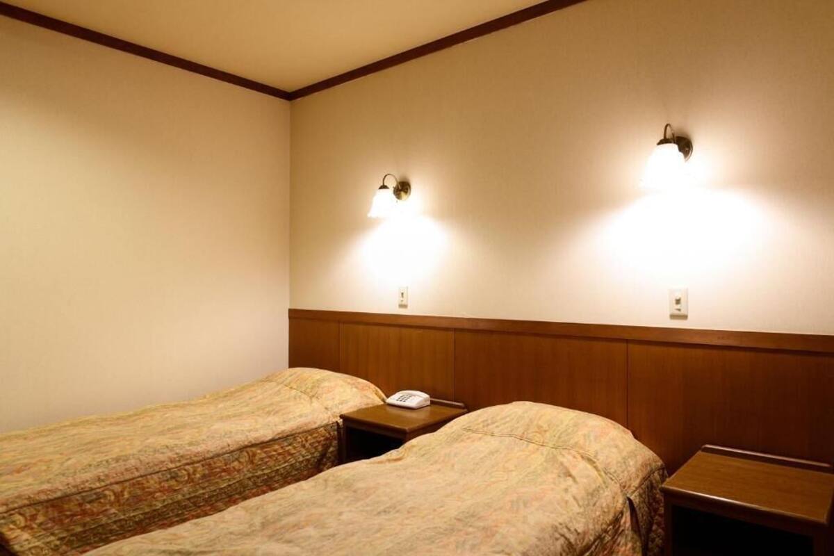 A hotel room with two single beds, wooden headboards, bedside tables, and wall-mounted lamps.
