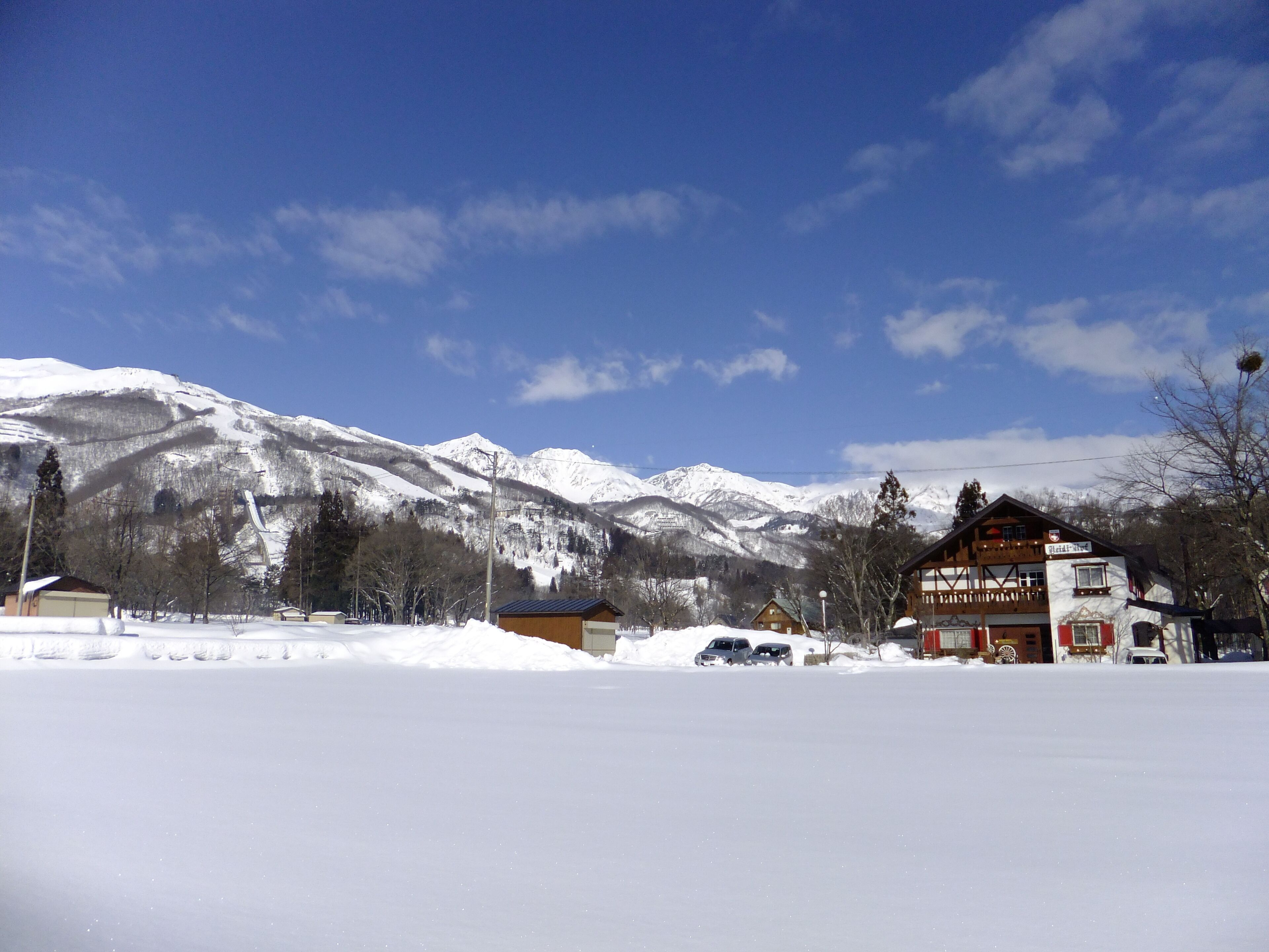 A snow-covered landscape with a mountain in the background, a building with a sign that reads "Hotel Alpenrose," and several parked cars.