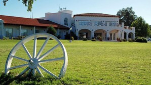 A large white building with a red roof and multiple arches in the front. A white wagon wheel is in the foreground on a grassy lawn. - La Campiña Club Hotel & Spa (Santa Rosa de Toay)