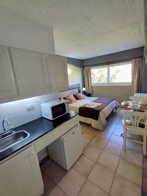 Une chambre d’hôtel compacte comprenant un coin cuisine, un lit recouvert d’une courtepointe marron, une fenêtre avec des rideaux et une petite table avec une lampe.