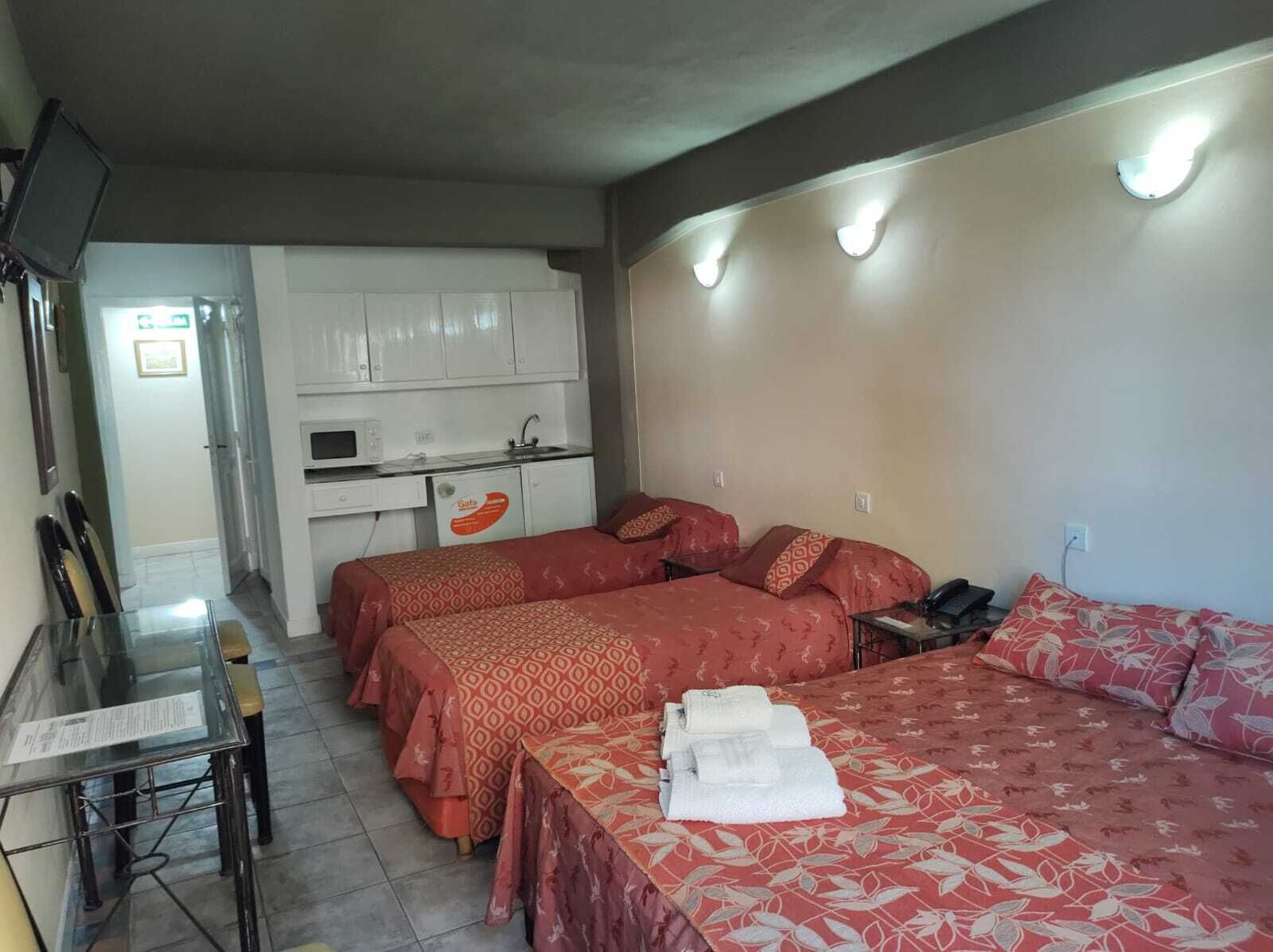 A compact hotel room with two beds, a small kitchenette, a glass table, and a TV.