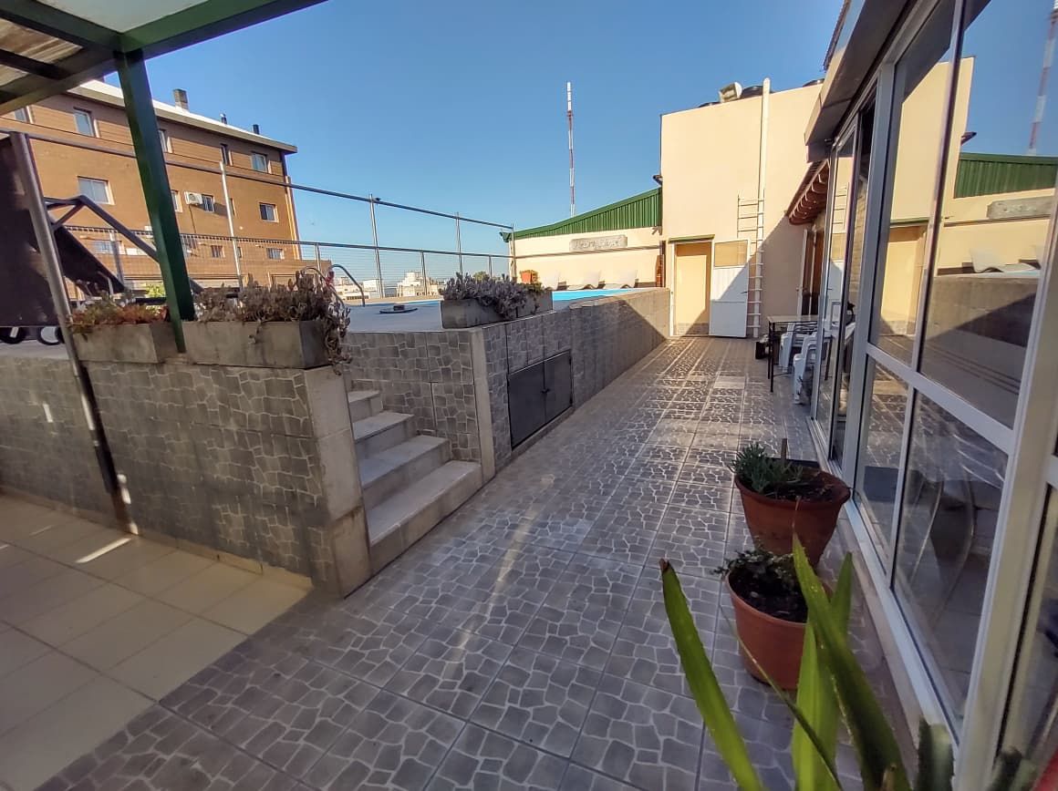 Terraço no telhado com piso de azulejos, plantas em vasos, escada e vista de prédios ao fundo.