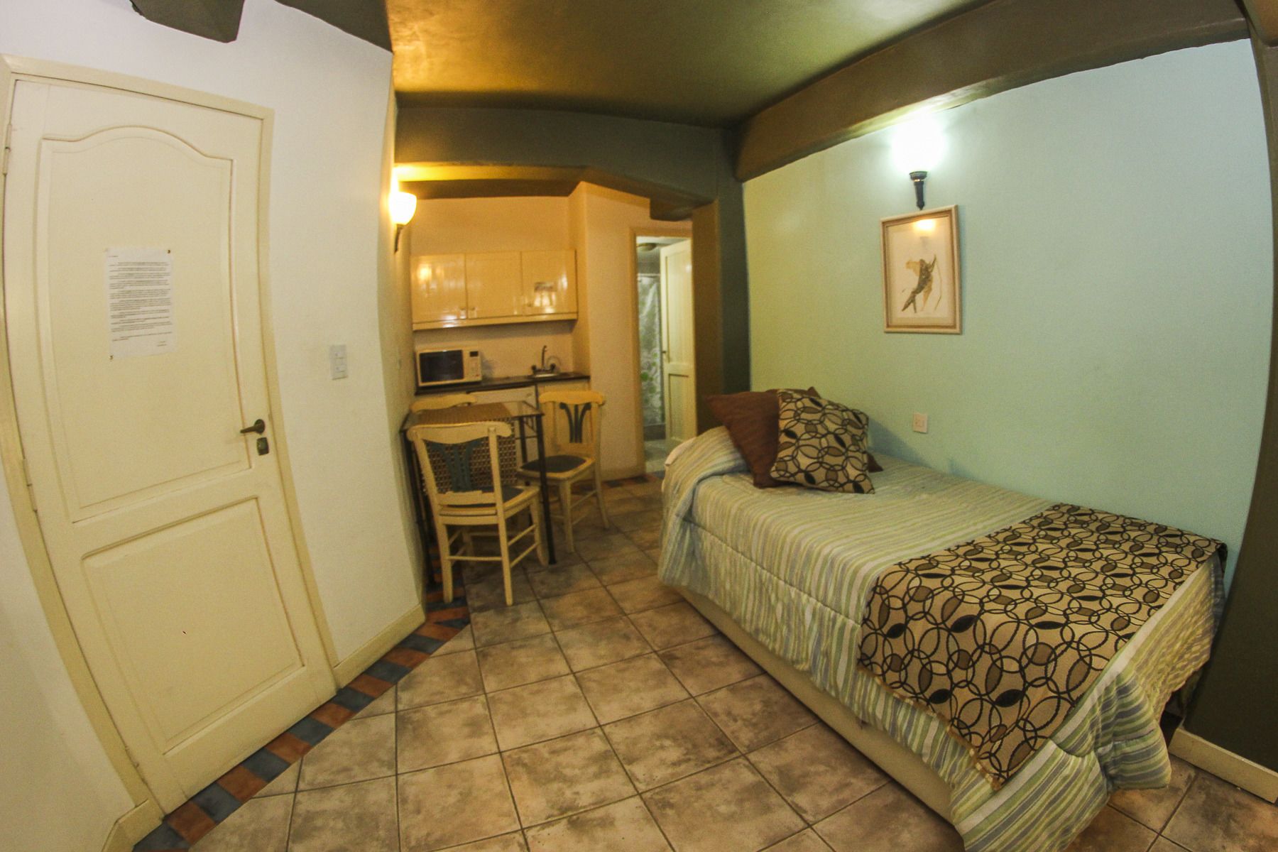 Quarto pequeno e compacto com cama, uma cadeira, mesa e cozinha compacta.