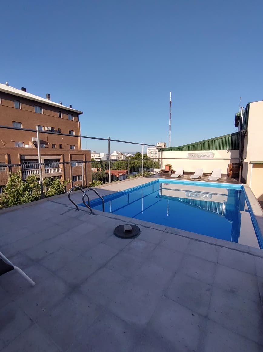 A rooftop swimming pool with a clear blue sky, a flagpole, and a building with a green roof in the background.