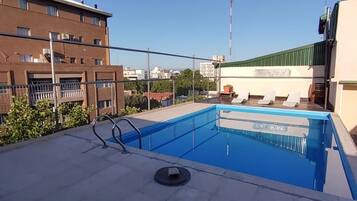 A rooftop swimming pool with a clear blue sky, a flagpole, and a building with a green roof in the background.