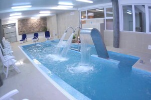 An indoor swimming pool with multiple water jets, white lounge chairs, and a stone wall in the background.