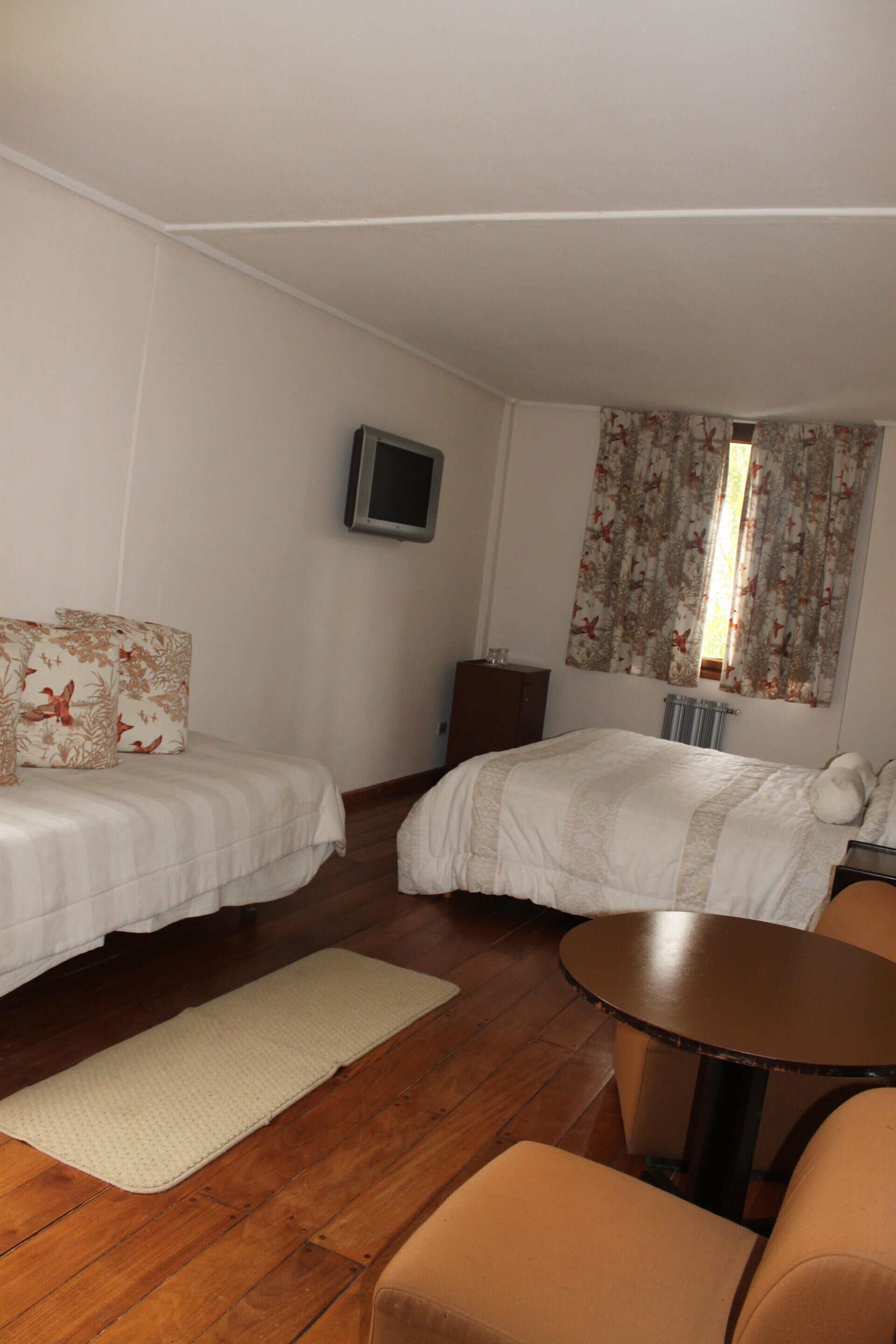 A bedroom with two beds, a TV, a window with curtains, a wooden floor, and a small round table.