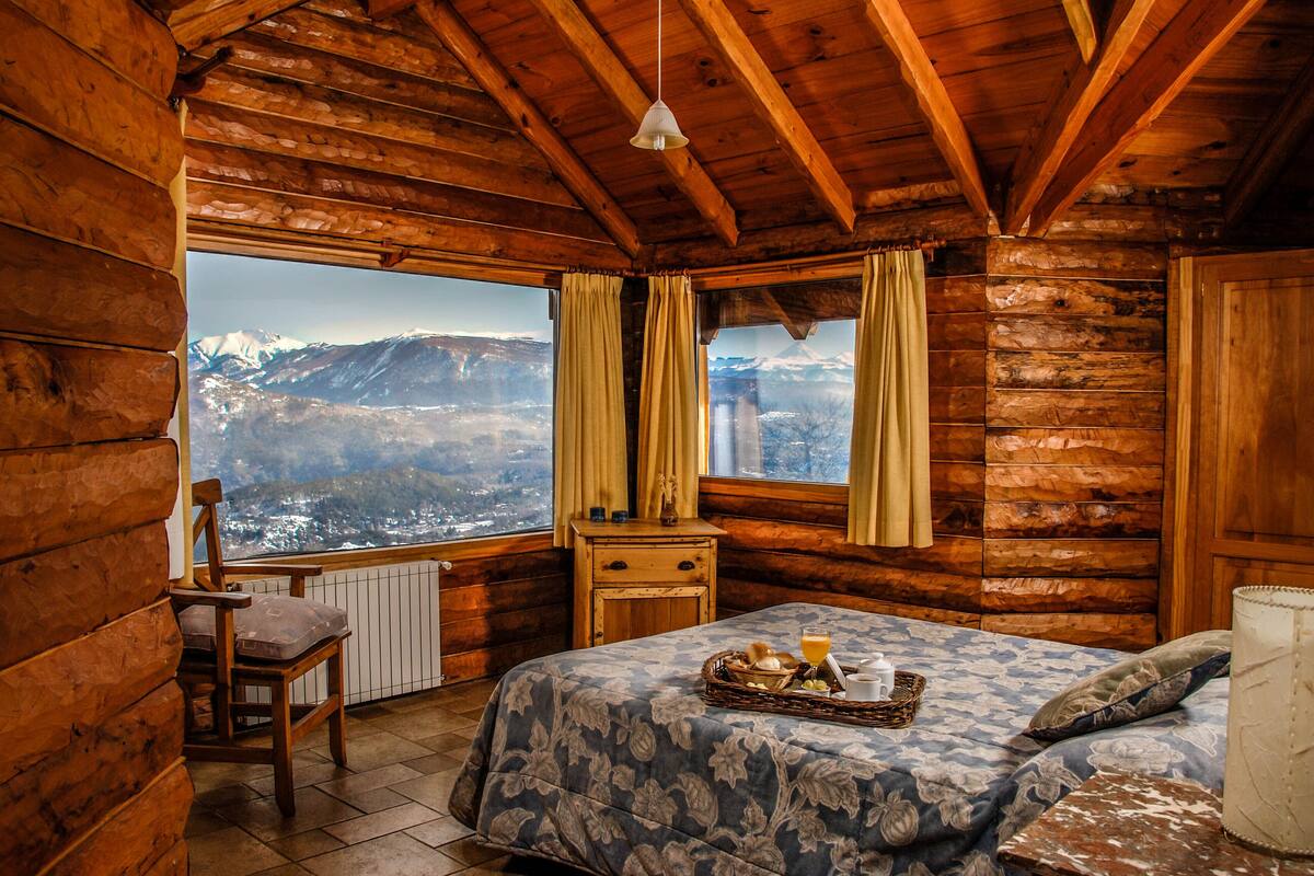 Una habitación tipo cabaña de madera con cama, una silla, una mesita de noche y una ventana con cortinas.