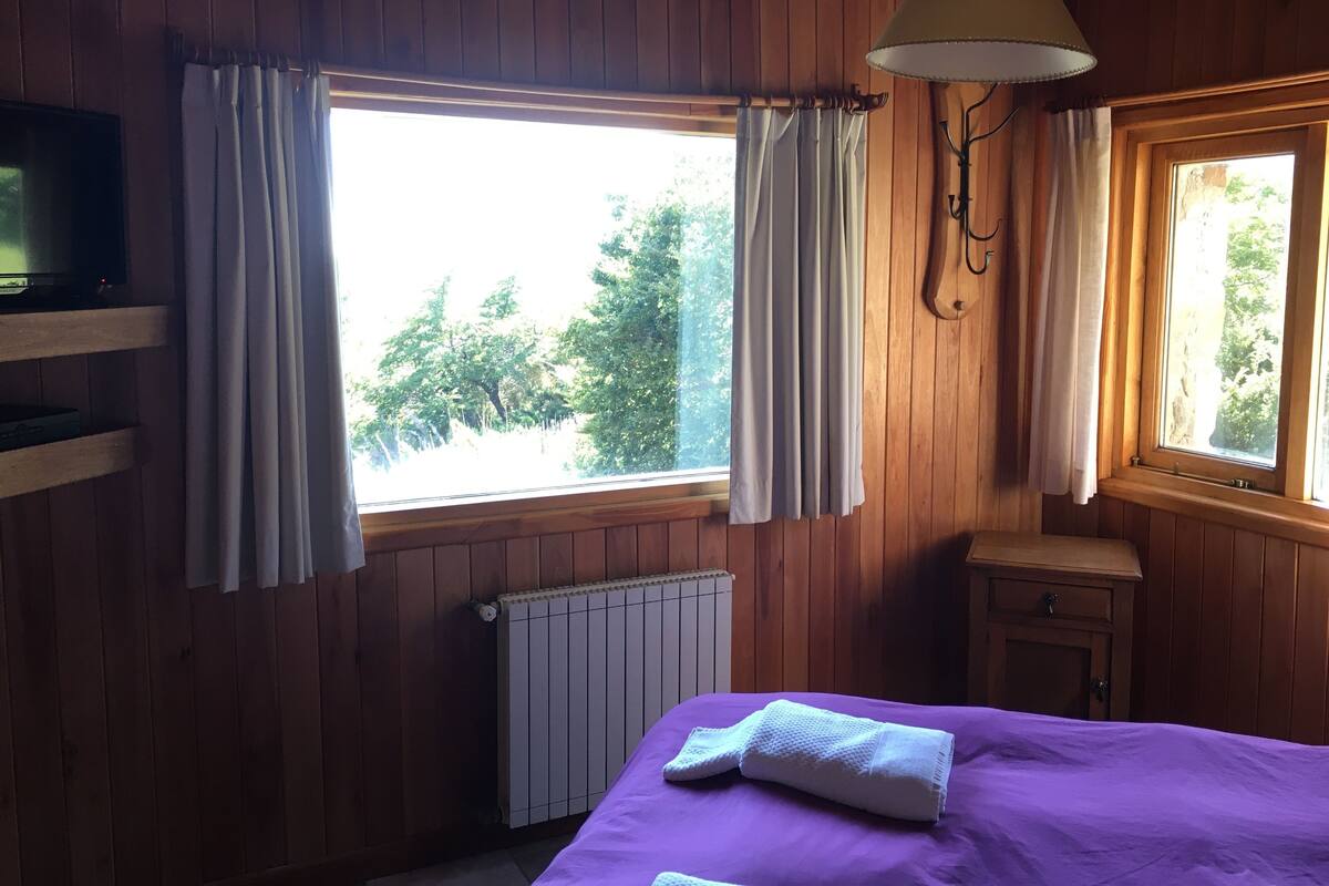 Una habitación con paredes de madera, una cama con colcha morada, un televisor sobre una estantería, una ventana con cortinas y un radiador.