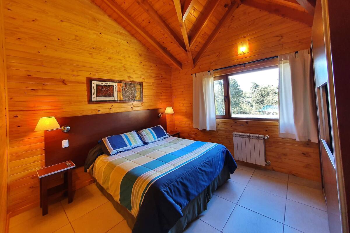 Un dormitorio de cabaña de madera con cama, mesitas de noche, ventana con cortinas y una lámpara fijada a la pared.