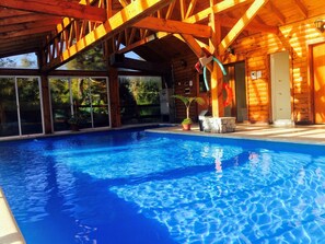 A wooden cabin with a swimming pool, large windows, and a stone fireplace.