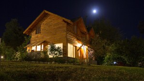 A wooden cabin with a stone foundation, illuminated by exterior lights, set against a night sky with a visible moon.