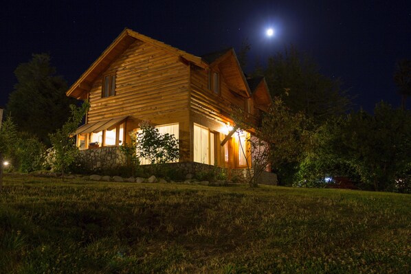 A wooden cabin with a stone foundation, illuminated by exterior lights, set against a night sky with a visible moon.