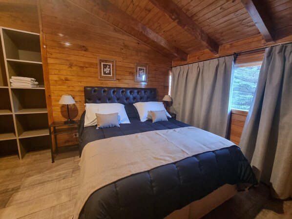 A wooden-paneled bedroom with a large bed, a nightstand, a shelf with folded towels, and two framed pictures on the wall.