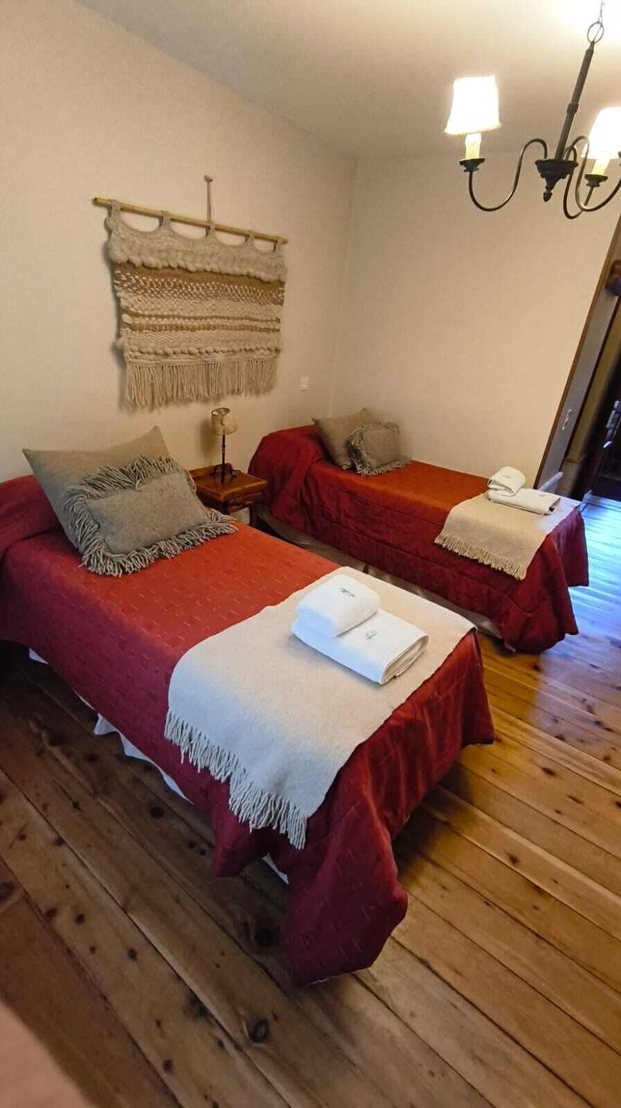 Ein Zimmer mit zwei Betten, Holzboden, Wandteppich, Wandlampe und einer Tür, die nach draußen führt.