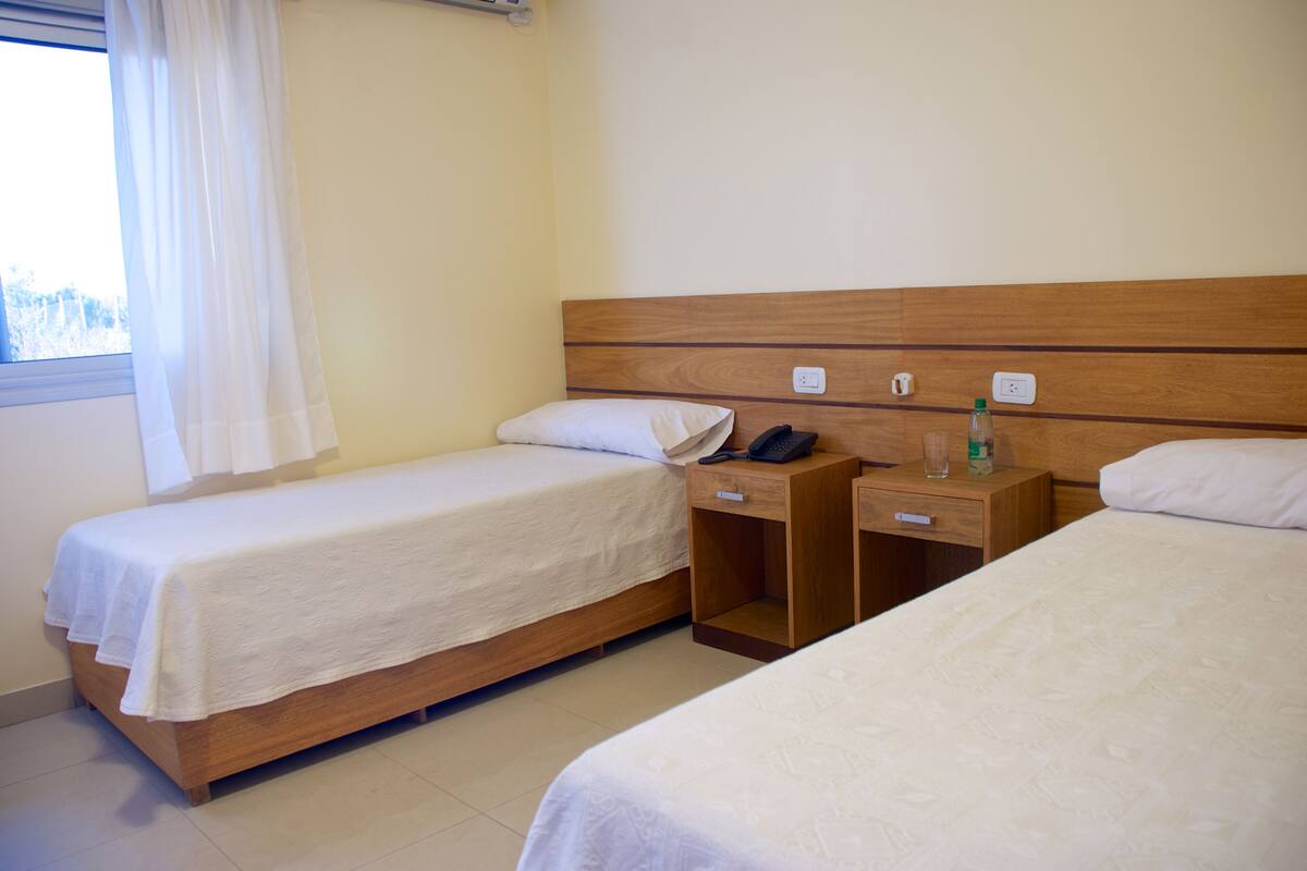 Ein Hotelzimmer mit zwei Einzelbetten, einem Holzkopfteil, Nachttischen mit Telefonen und Pflegeartikeln sowie einem Fenster mit Vorhängen.