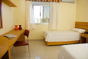 Un dormitorio con una cama individual, un escritorio de madera con una computadora portátil, dos jarrones rojos y una ventana con cortinas.