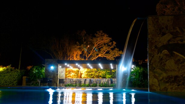 Una piscina iluminada con luz ambiental, con caída de agua y rodeada de mobiliario de exterior.