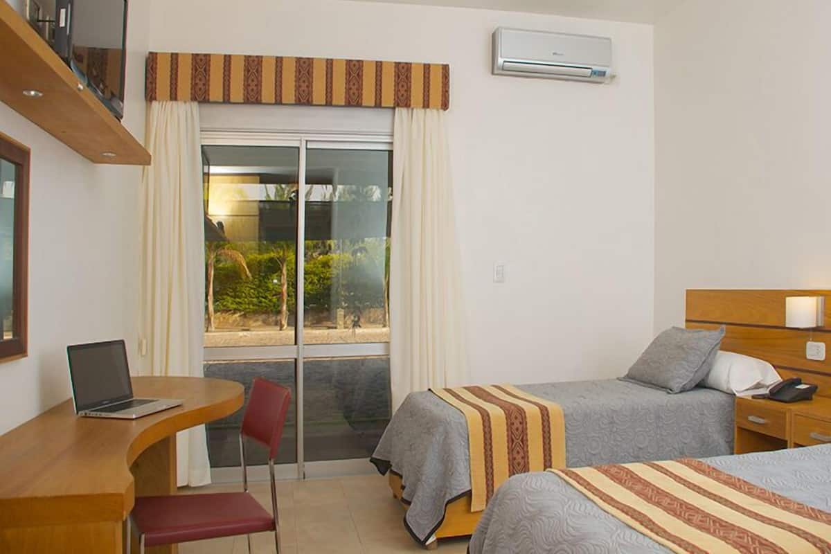 Ein Hotelzimmer mit zwei Betten, einem Schreibtisch mit Laptop, einem Sessel, einem Fernseher auf einem Regal und einer Klimaanlage.