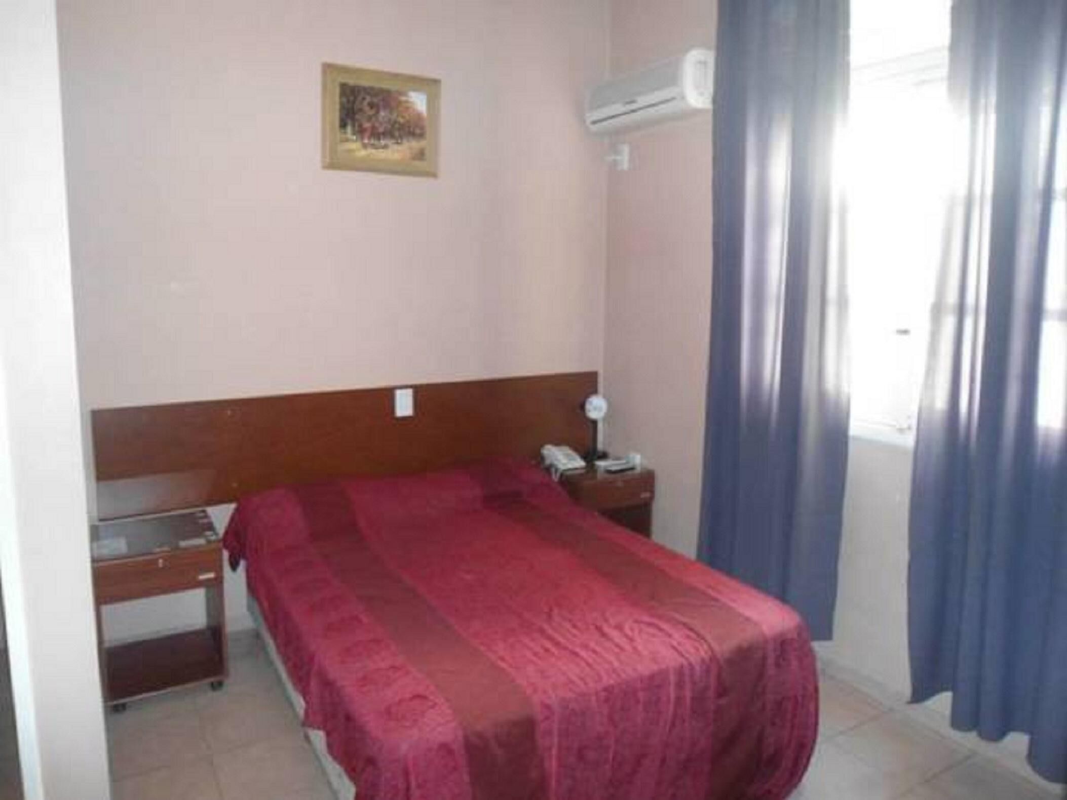 A hotel room with a single bed, a bedside table with a lamp, a small desk with a chair, a wall-mounted air conditioner, and a window with curtains.
