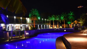 Uma área de piscina noturna com design curvo, palmeiras e um edifício com janelas iluminadas.