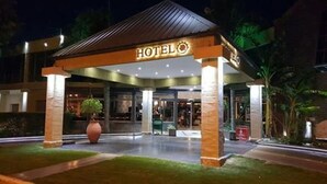 Entrada de hotel com área coberta, um vaso grande com planta e fachada bem iluminada.