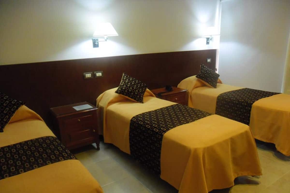 Une chambre d’hôtel avec quatre lits, chacun agrémenté d’une couvre-lit et de coussins à motifs, une table de chevet avec une lampe et un luminaire fixé au mur.