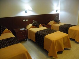 Habitación de hotel con cuatro camas, cada una con una colcha y almohadas estampadas, una mesita de noche con lámpara, y una lámpara de pared.