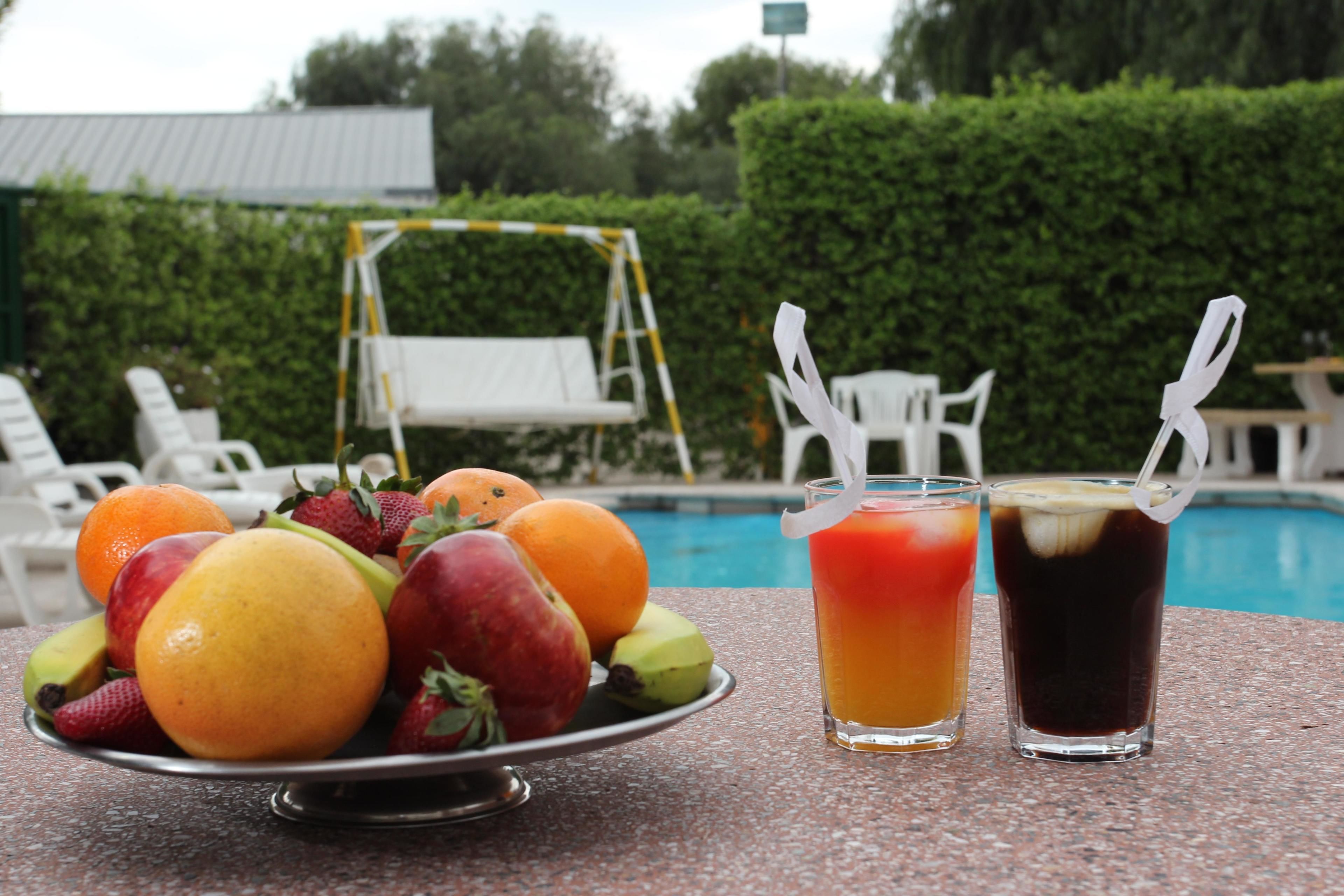 Una mesa con una bandeja de frutas y dos bebidas, una piscina y sillones de descanso al fondo.