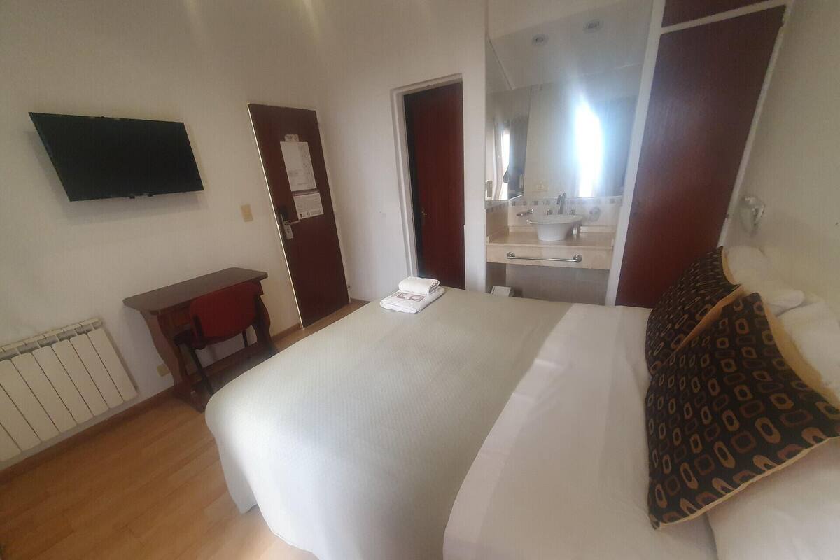 Une chambre d’hôtel avec un lit, un bureau, une chaise, un téléviseur fixé au mur et une salle de bain visible à travers une porte ouverte.