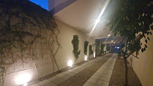 Un couloir éclairé la nuit, avec un sol carrelé, un mur recouvert de plantes grimpantes et un arbre sur la droite.