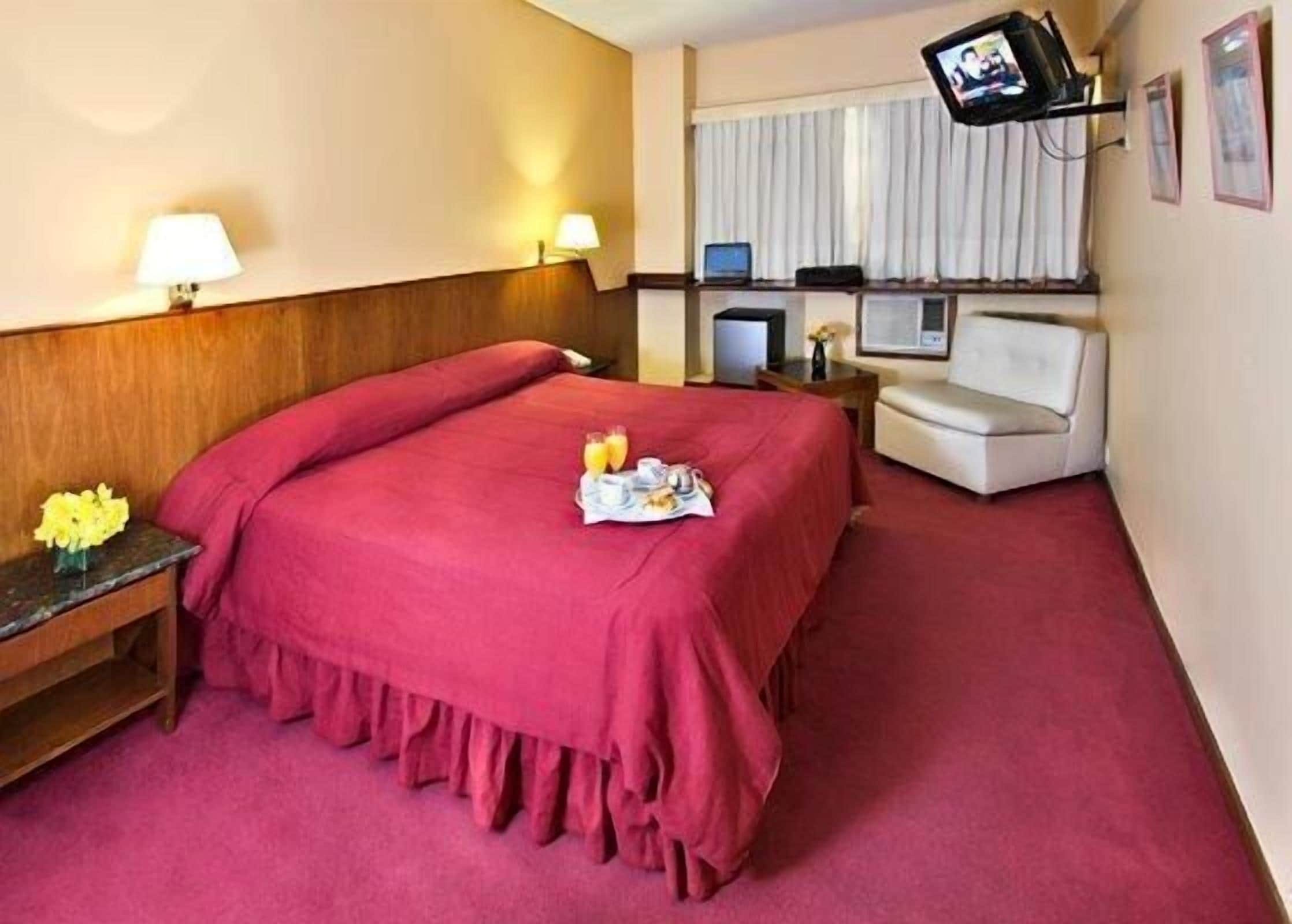 A hotel room with a large bed, a desk with a computer, a TV, and a small table with flowers.