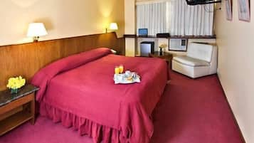 Habitación de hotel con una cama grande, un escritorio con computadora, un televisor y una mesita con flores.