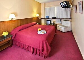 Quarto de hotel com uma cama grande, uma escrivaninha com um computador, uma televisão e uma pequena mesa com flores.