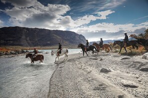 Um grupo de pessoas a cavalo ao longo de uma margem de rio, com montanhas ao fundo.