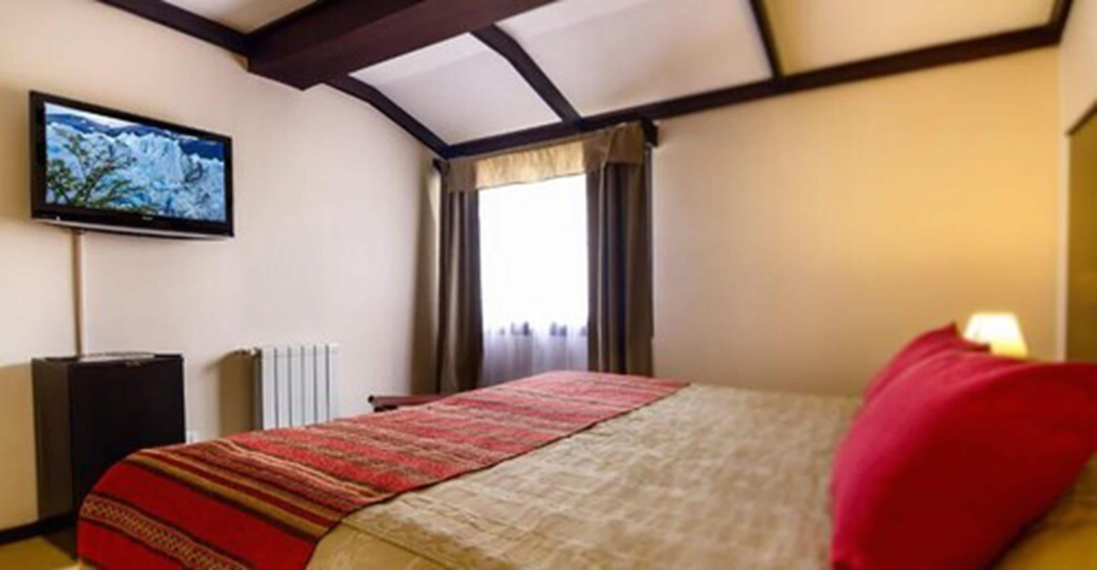 A hotel room with a bed, a television on the wall, and a window with curtains.