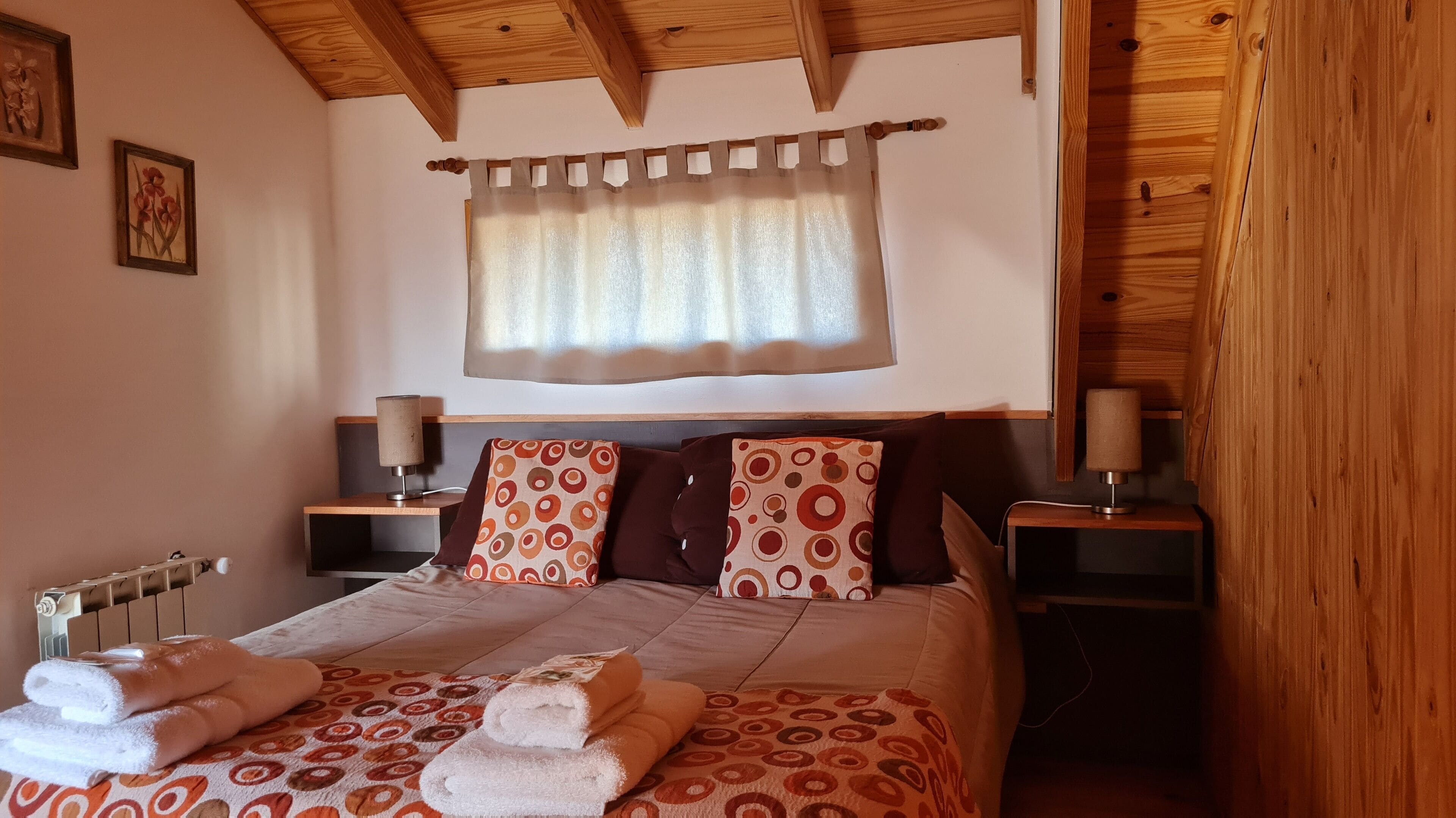 A bedroom with a wooden ceiling, a bed with patterned bedding, two bedside tables with lamps, and a window with a curtain.