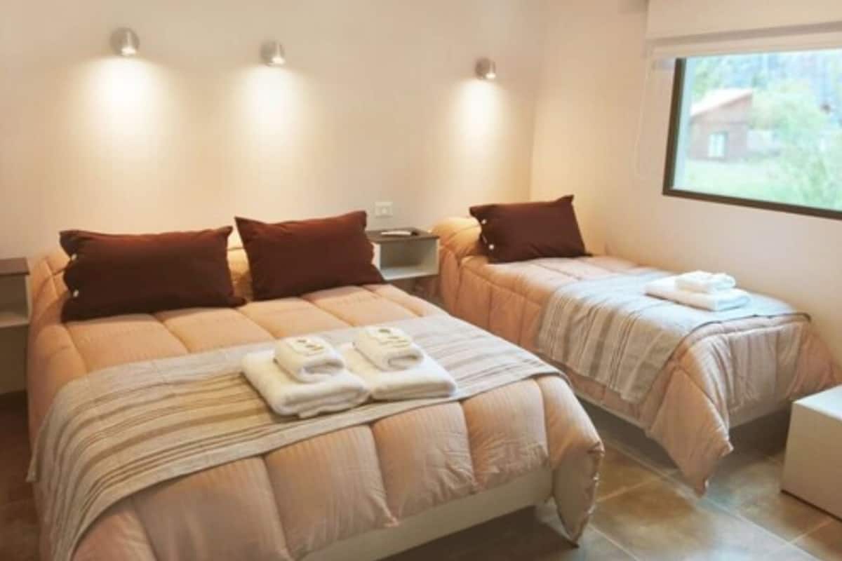 Habitación de hotel con dos camas, cada una con una almohada marrón y una toalla doblada. Hay luces fijadas a la pared y una ventana con vistas a la vegetación.