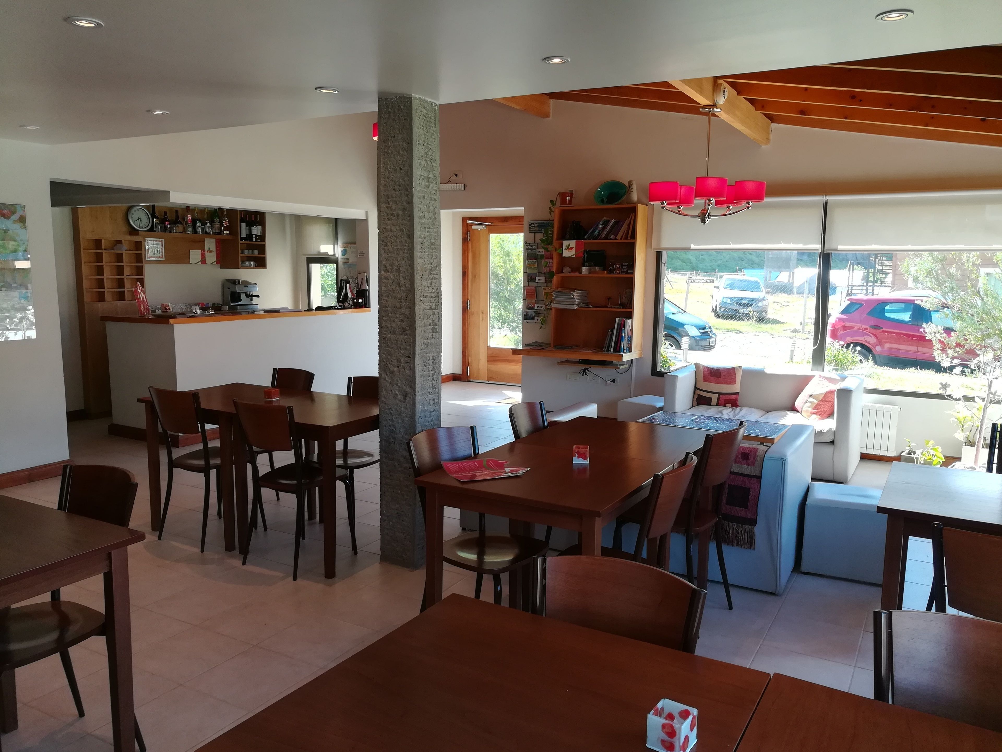 A modern dining area with wooden tables and chairs, a kitchen area with a bar, and a view of parked cars outside.
