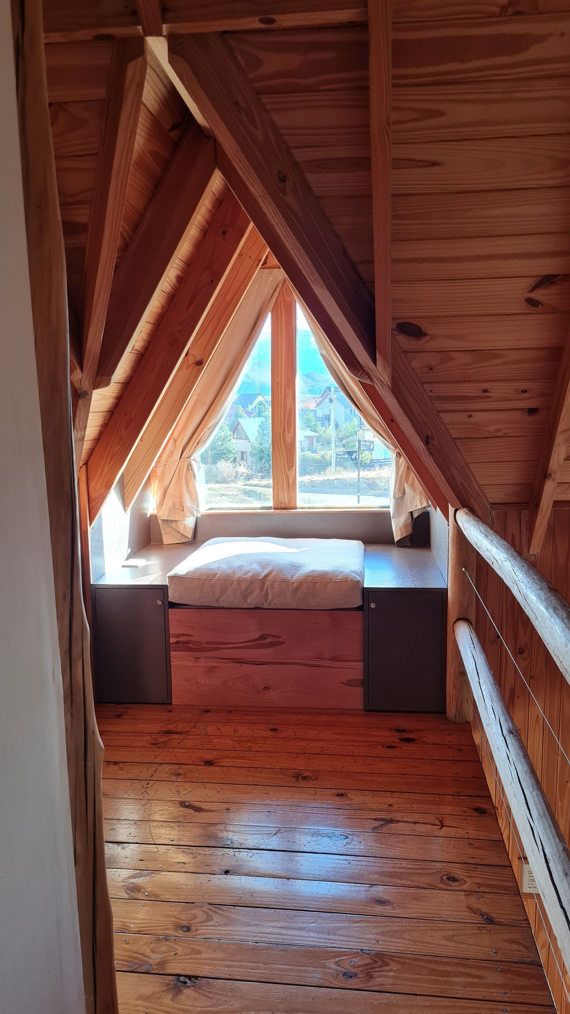 Ett sovrum med ett sluttande tak, synliga träbjälkar, ett fönster med utsikt över naturen, en säng med en kudde och ett trägolv.