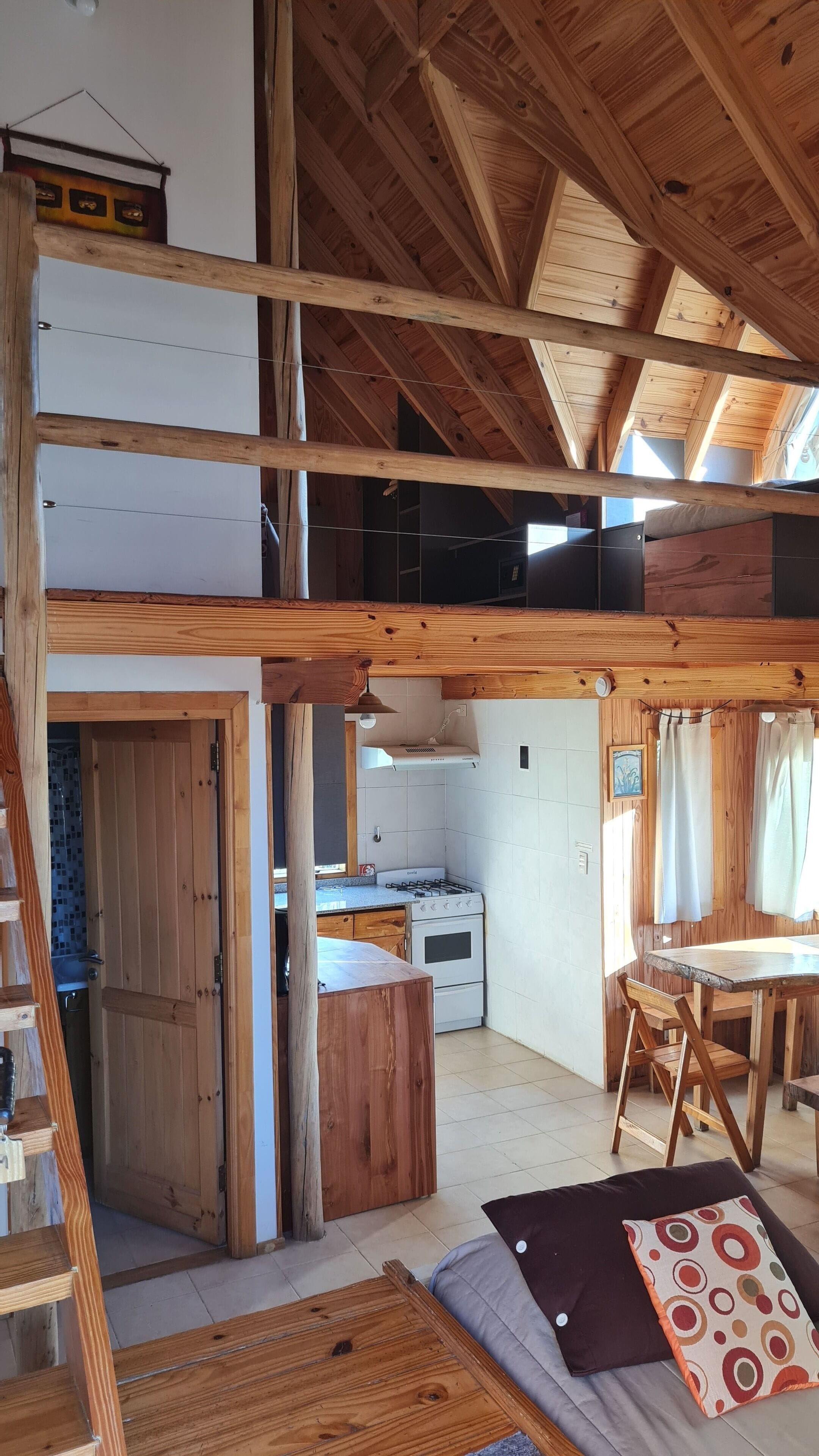 A rustic cabin interior with a loft bedroom, wooden beams, a kitchen area, and a dining space.