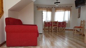 A living room with a red sofa, wooden dining table with chairs, and a television mounted on the wall. - Los Pinos Apart Hotel (Ushuaia)