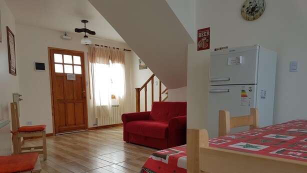 A cozy room with a wooden floor, a red sofa, a wooden chair, a small table, a bed with a Christmas-themed bedspread, a white refrigerator, a clock, and a door with a window. - Los Pinos Apart Hotel (Ushuaia)