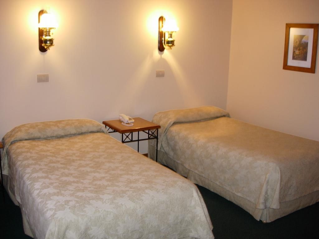 Habitación de hotel con dos camas individuales, una mesita pequeña y dos lámparas fijadas a la pared.