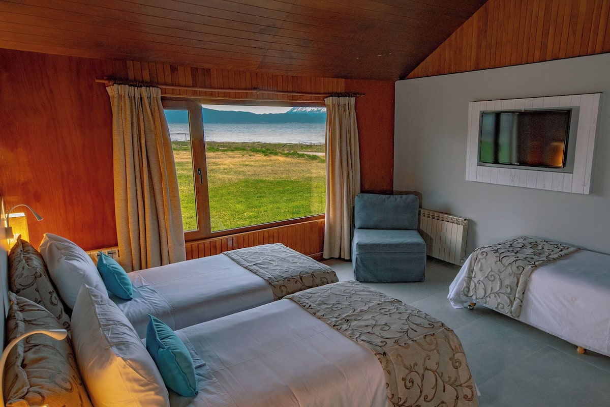 A room with two beds, a chair, and a window with a view of a field and mountains.