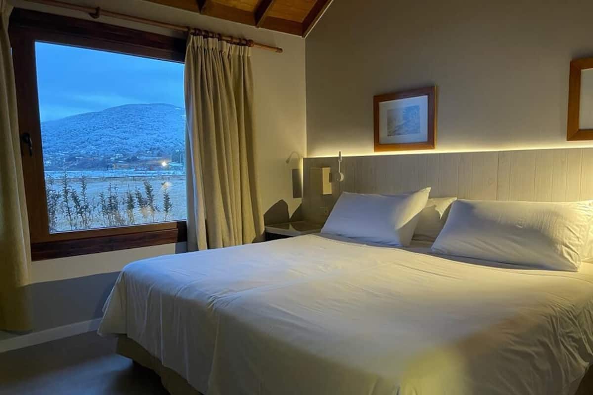 A bedroom with a large bed, a nightstand, a bedside table, a window with curtains, and a framed picture on the wall.