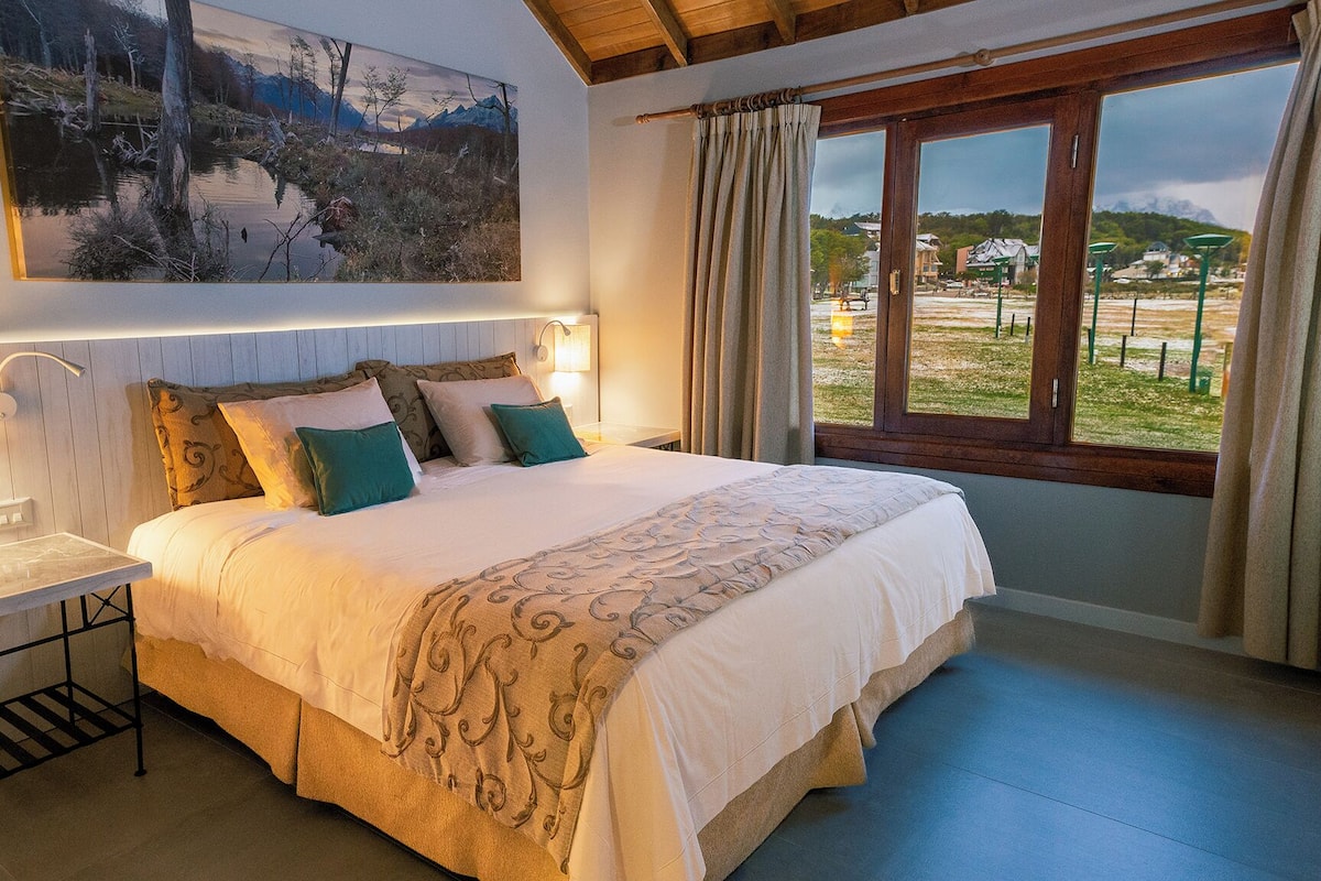 A bedroom with a large bed, bedside lamps, a painting, and a view of the outdoors.